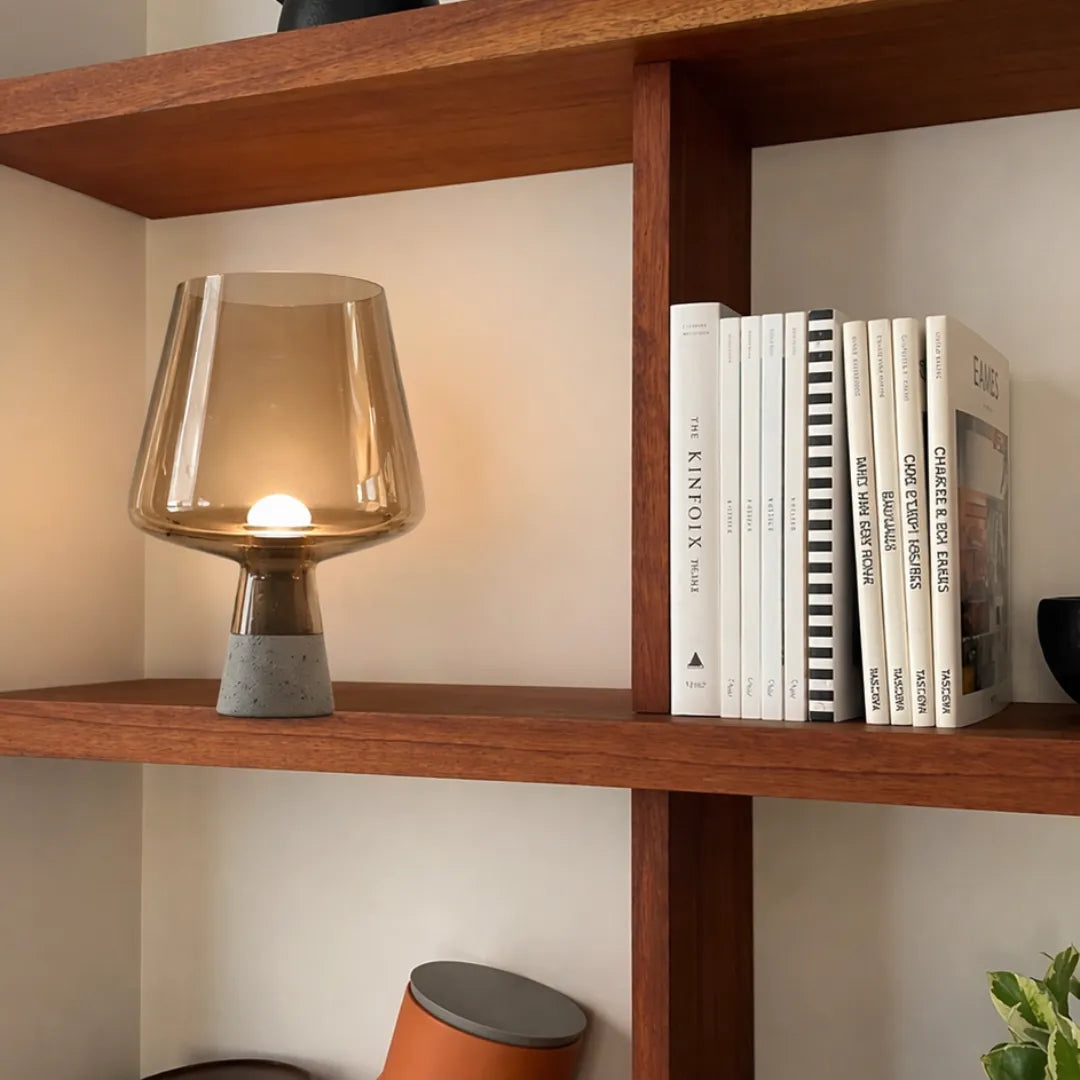 Luminária de mesa em vidro âmbar acesa em prateleira de madeira ao lado de pilha de livros brancos em estante moderna — Luminária de Mesa Moderna Vidro Base Mármore — Tonzi