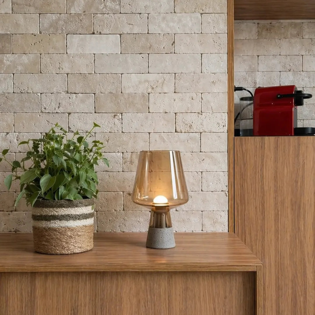 Luminária de mesa em vidro âmbar acesa sobre balcão de madeira em frente a parede de tijolos travertino com vaso de planta verde — Luminária de Mesa Moderna Vidro Base Mármore — Tonzi