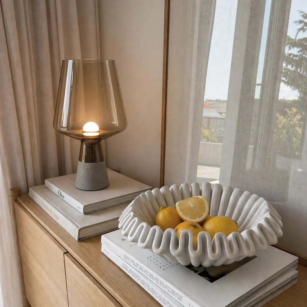 Luminária de mesa em vidro âmbar acesa sobre pilha de livros em console de madeira ao lado de centro de mesa cerâmico com limões e janela com cortina translucida — Luminária de Mesa Moderna Vidro Base Mármore — Tonzi