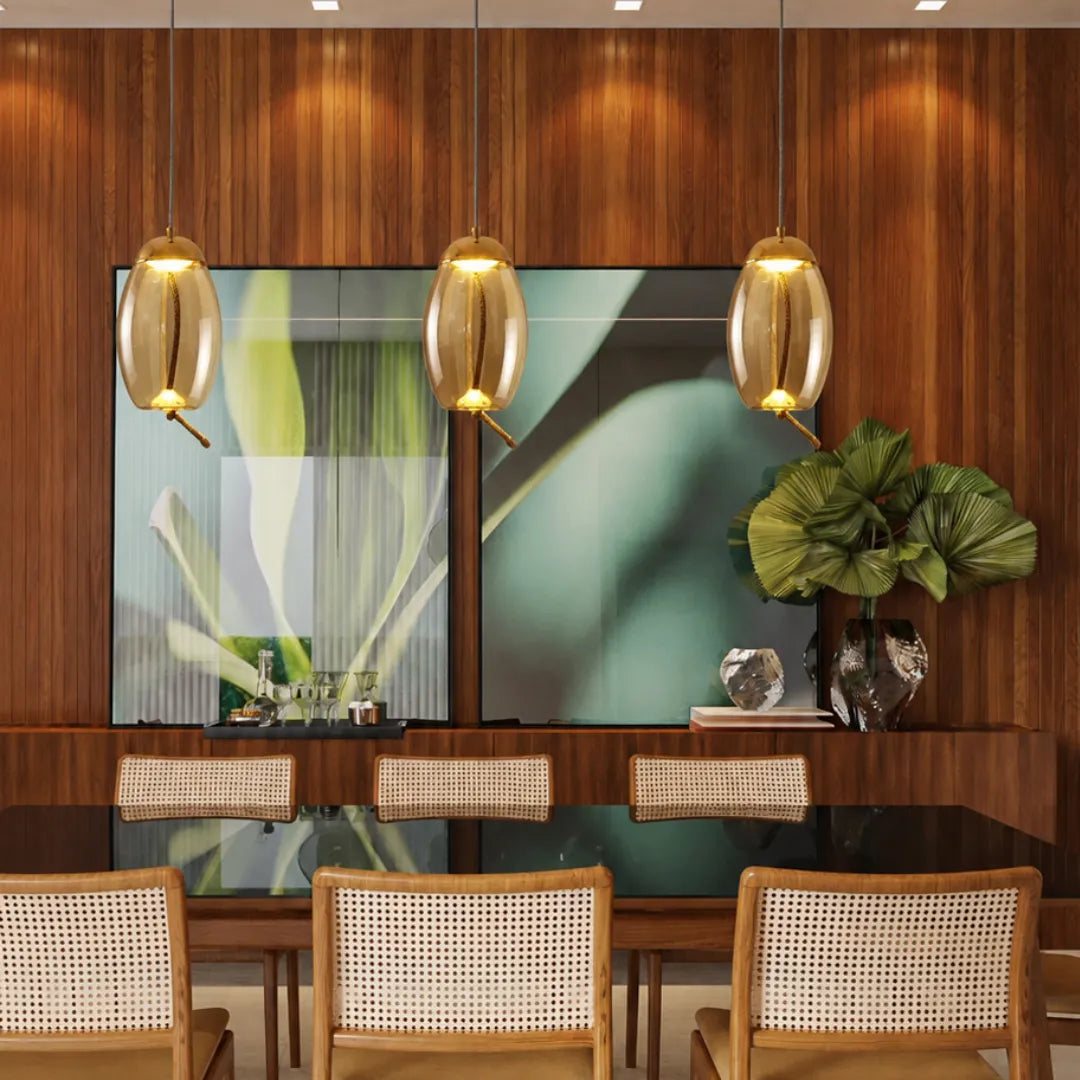 Trio de pendentes ovais âmbar Cognac sobre mesa de jantar com painel de madeira e cadeiras de palhinha — Luminária Pendente Vidro Soprado LED Minimalista Névoa — Tonzi